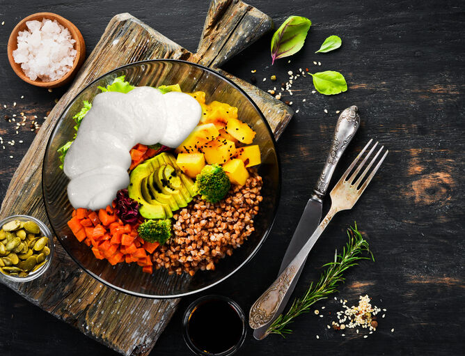 Bowl mit Mozzarella von GOLDSTEIG, Avocado, Paprika, Salat und Brokkoli in Glasschale auf Holzbrett, daneben Besteck