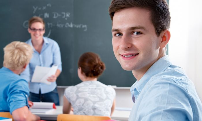 Schüler: Ein Schüler blickt sitzend im Klassenzimmer über seine Schulter in die Kamera. Im Hintergrund sieht man seine Schulkameraden.