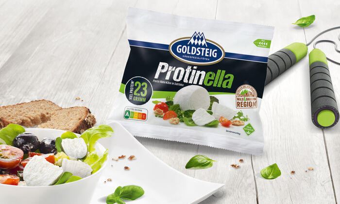 Protinella von GOLDSTEIG, der Mozzarella mit viel Eiweiß. Verpackung drapiert auf grauem Holztisch mit einem leckeren Salat und einem Springseil.