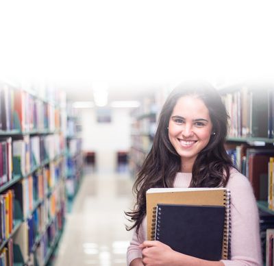 Infos zur Bewerbung: Studentin hält ihre Bücher in der Hand und blickt in die Kamera. Im Hintergrund sieht man die Bibliothek.