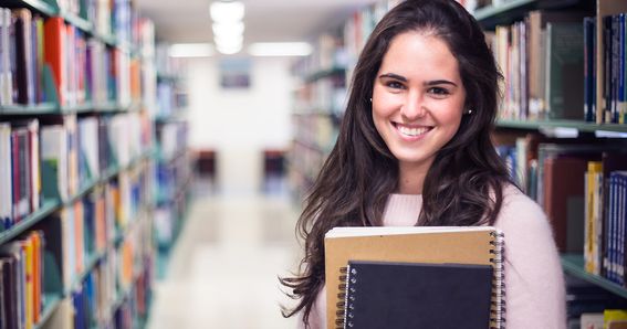 Karriere: Studentin hält ihre Bücher in der Hand und blickt in die Kamera. Im Hintergrund sieht man die Bibliothek.