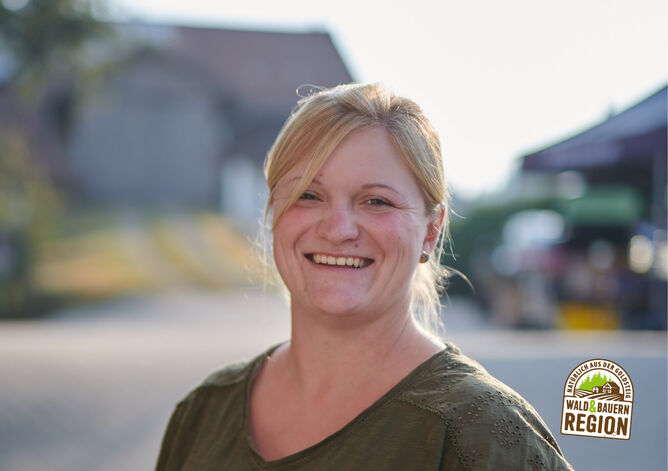 Startseite: Landwirtin, die zu den GOLDSTEIG Käsereien liefert, lächelt in die Kamera. Im Hintergrund kann man verschwommen ihren Bauernhof erkennen. Unten rechts ist das Herkunftssiegel "Natürlich aus der GOLDSTEIG Wald & Bauernregion" zu sehen.