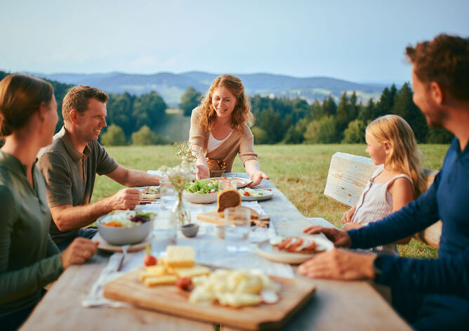 Startseite: Urlaub auf dem Bauernhof. Die Familie und das Urlauber Pärchen machen zusammen draußen Brotzeit. Die Landwirtin stellt einen leckeren Caprese Teller mit GOLDSTEIG Mozzarella auf den Tisch.
