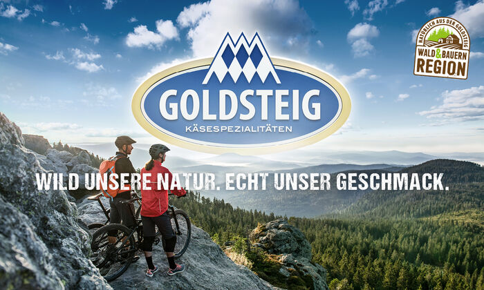 Protinella (Proteinkäse): Pärchen in Sportklamotten sehen am Rande eines Berges am Arber in die Weite des Bayerischen Waldes und halten dabei deren Mountainbikes. Oben rechts das Herkunftssiegel "Natürlich aus der GOLDSTEIG Wald & Bayernregion", mittig das GOLDSTEIG-Logo und darunter der Slogan "Wild unsere Natur. Echt unser Geschmack."