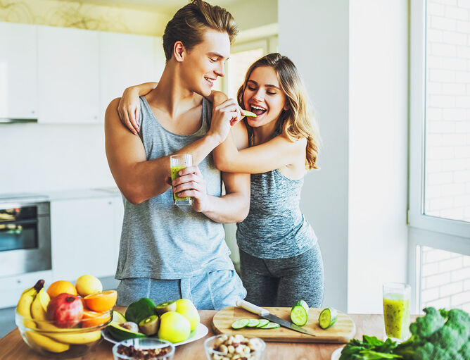 Pärchen in Sportkleidung, Mann gibt Frau Apfelspalte und hält grünen Smoothie in der Hand, auf dem Esstisch liegt Obst, Gemüse und Nüsse 