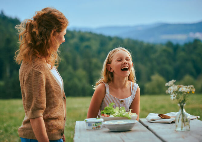 Startseite: Landwirtin, die zu den GOLDSTEIG Käsereien liefert, sieht ihre Tochter an. Ihre Tochter lacht herzhaft. Auf dem Tisch ist ein Salat mit GOLDSTEIG Bambini Mozzarella Mini zu sehen.