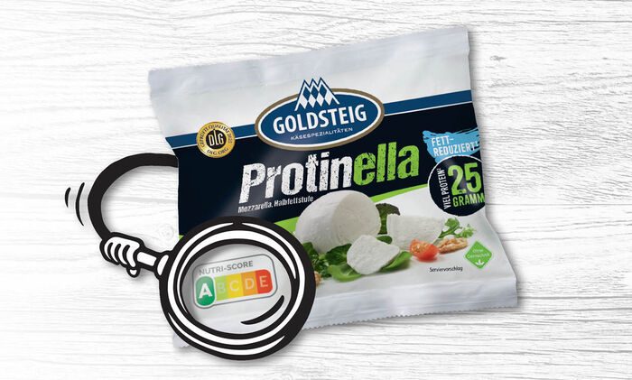 All about Nutri-Score/products: White background with a wooden look. On it you can see a GOLDSTEIG Protinella ball packaging with an illustrated arm, which emphasises the Nutri-Score A on the packaging with a magnifying glass.