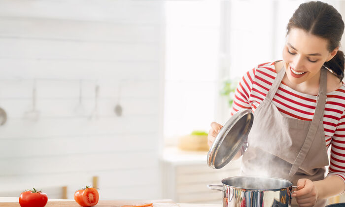 Frau steht mit Schürze in der Küche und hebt gerade den Topfdeckel an. Auf der Arbeitsfläche sieht man geschnittene Tomaten liegen.
