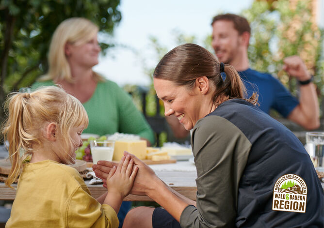 Startseite: Urlaub auf dem Bauernhof. Die Familie und das Urlauber Pärchen machen zusammen draußen Brotzeit. Die Landwirtin unterhält sich mit dem Urlauber und das Kind der Landwirtin spielt mit der Urlauberin ein Klatschspiel. Unten rechts ist das Herkunftssiegel "Natürlich aus der GOLDSTEIG Wald & Bauernregion" zu sehen.